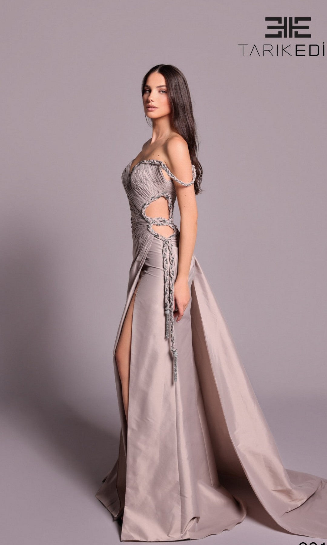 A woman stands in profile wearing the Tarik Ediz 99108 long formal dress in mink, featuring an off-shoulder sweetheart neckline, side cutouts, draping, and a long train. The background is plain light gray.